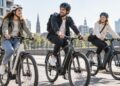 Drei junge Erwachsene fahren mit Helmen auf modernen Urban-E-Bikes durch eine helle europäische Stadt.